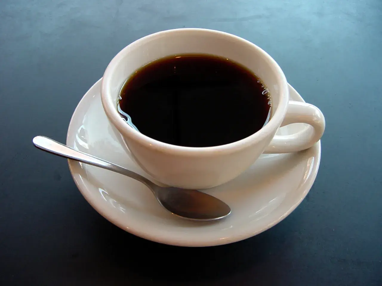 En vit kopp med mörkt kaffe och en sked på en fat. En påminnelse om kaffe ursprung och dess resa till din kopp.