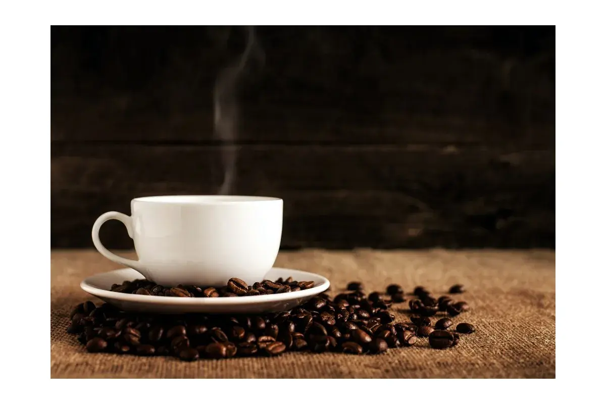 En vit kaffekopp med ånga stiger från den, omgiven av kaffebönor. En perfekt bild för att fundera på hur många skopor kaffe till 2 koppar.