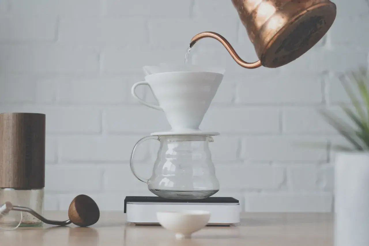 Vatten hälls från en kopparkanna över ett filter i en vit keramiktratt, som står på en glaskaraff. En kaffekvarn och en träsked ligger bredvid. Det är dags att brygg.