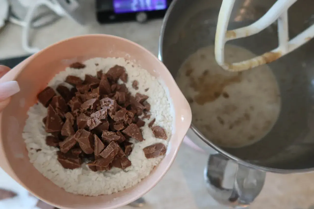En skål med mjöl och chokladbitar för en saftig banankaka med choklad. Bredvid står en köksmaskin med smet.