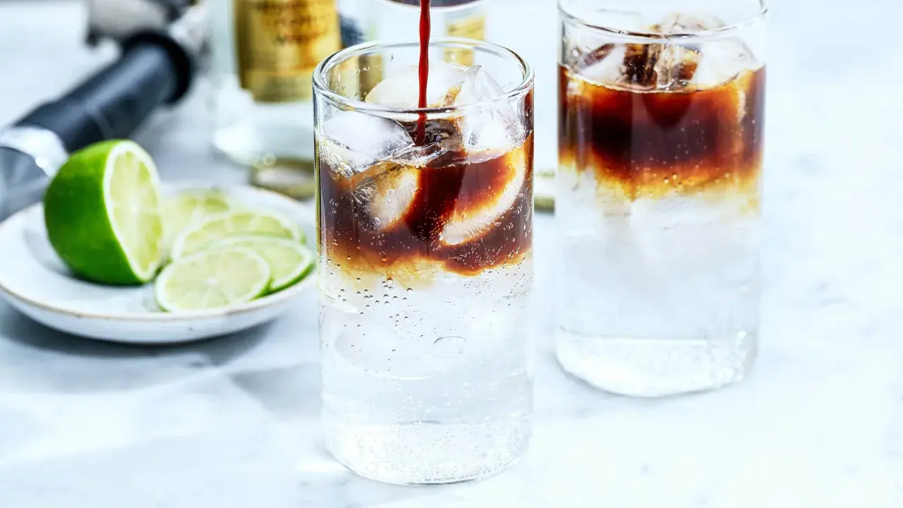 Två glas med kaffe tonic, is och lime. En flaska och en limeklyfta i bakgrunden.