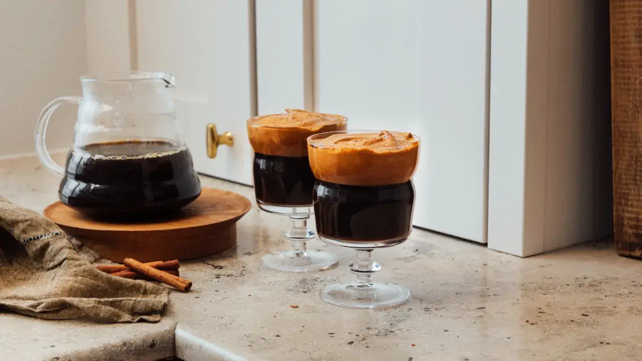 Två glas med mörkt kaffe toppat med fluffigt skum, en härlig dalgona kaffe-upplevelse. Bredvid står en karaff med kaffe och kanelstänger.
