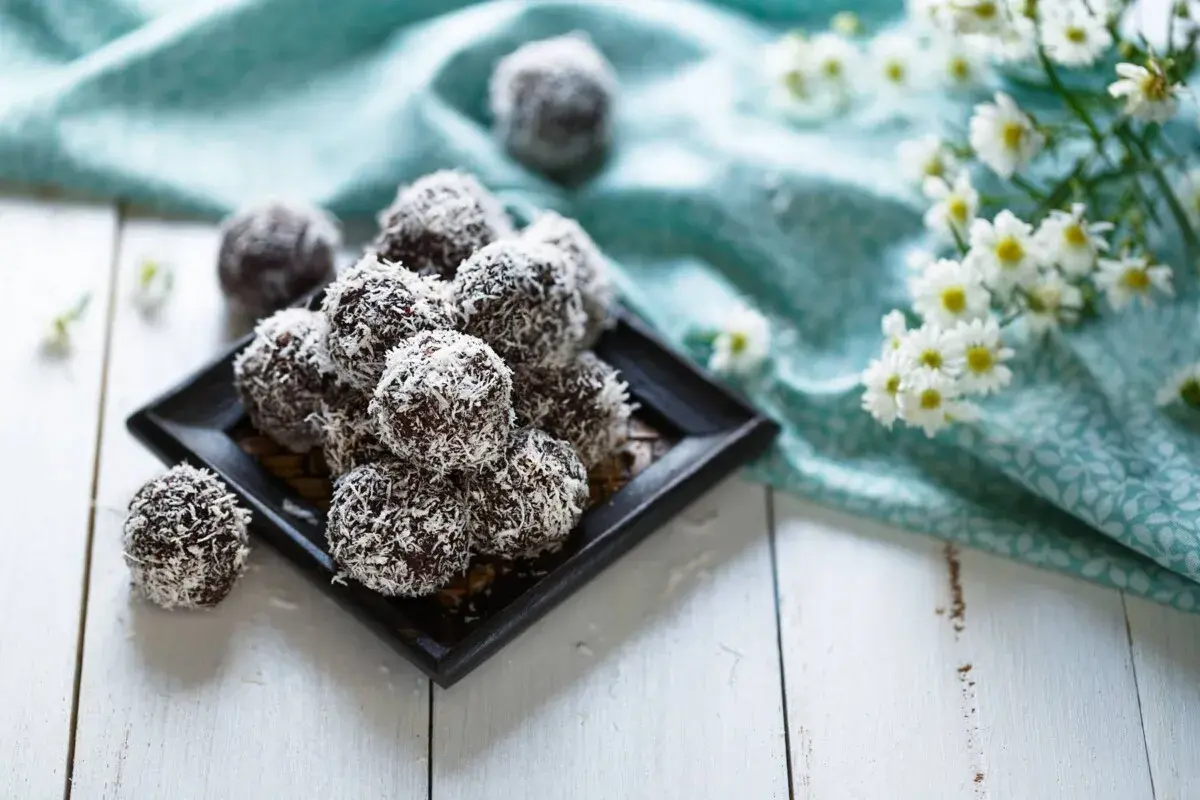 En hög med chokladbollar, perfekta för chokladbollens dag, rullade i kokosflingor. Blommor och en turkos duk i bakgrunden.