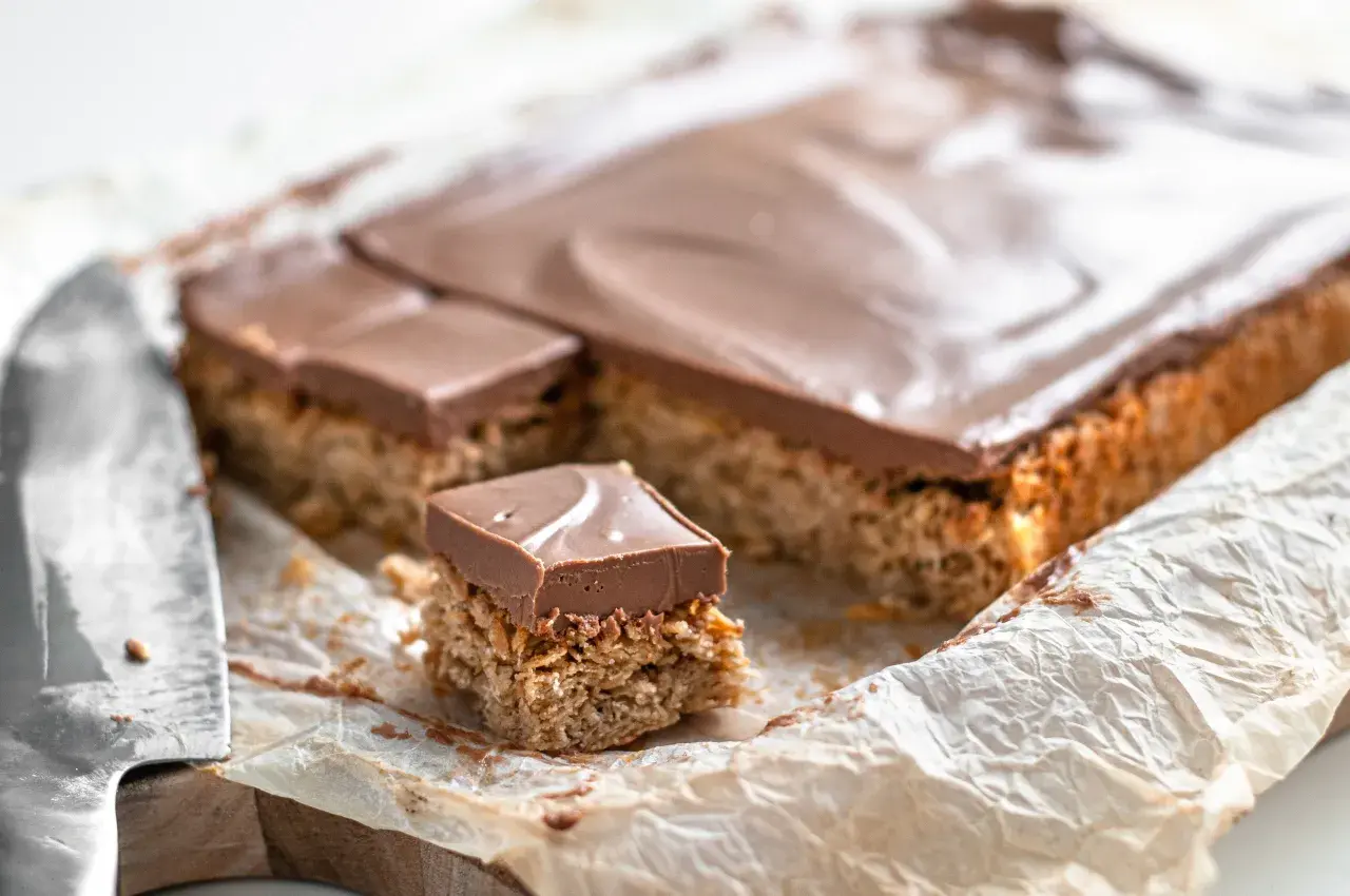 En bit av en kaka med chokladöverdrag, som ser ut att vara nougatrutor glutenfria, ligger på bakplåtspapper bredvid en kniv.