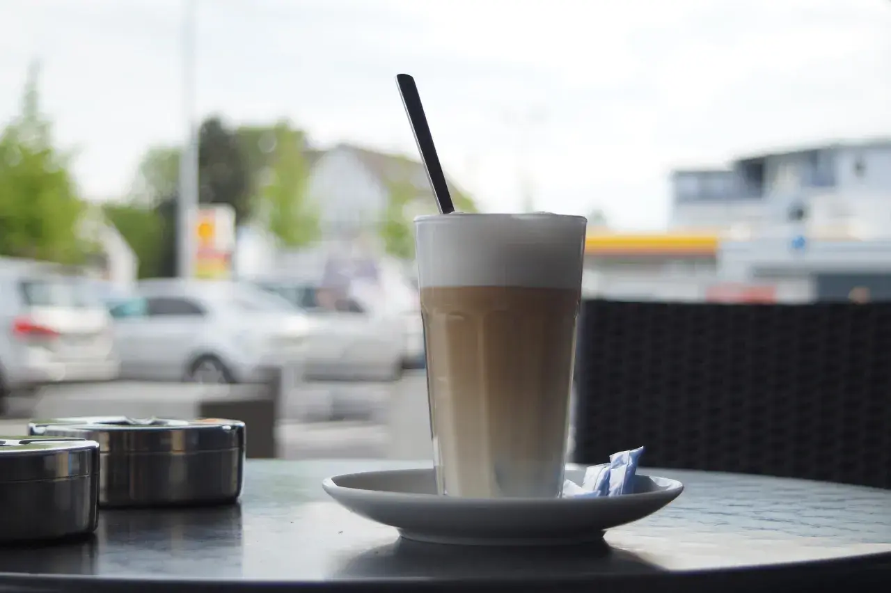 En hög latte macchiato med skummjölk och en sked på ett fat, bredvid två askkoppar.