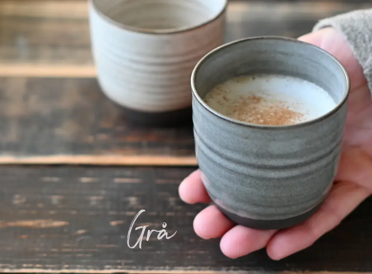 En hand håller en grå mugg med ett skummande, kaneldoftande lungo kaffe. En annan mugg syns i bakgrunden.