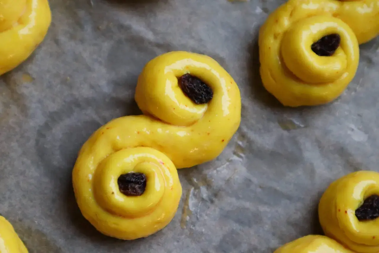 Gyllene, S-formade glutenfria bullar med russin på bakplåtspapper. Perfekt för fikat!