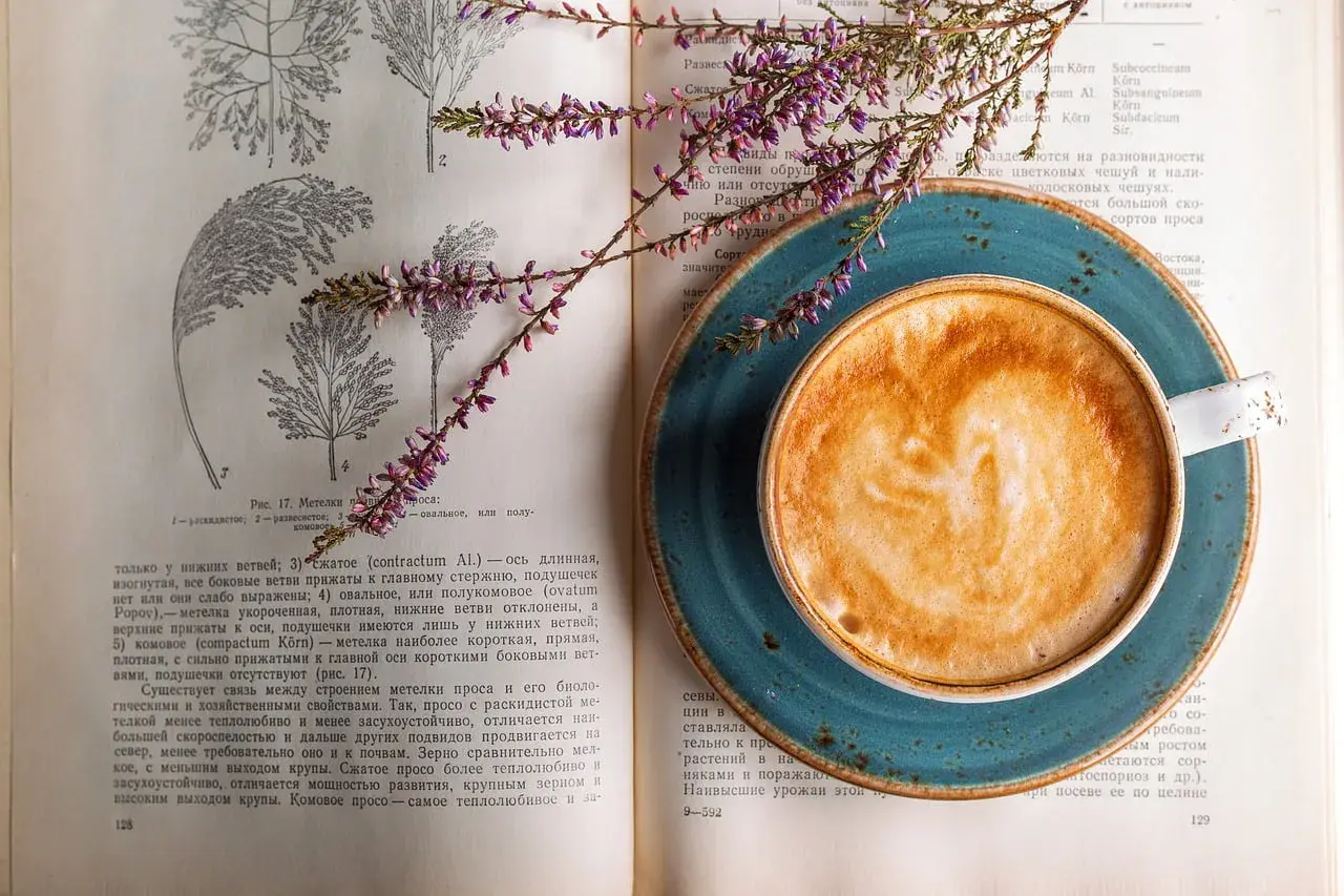 En kopp kaffe med lattekonst vilar på en öppen bok med botaniska illustrationer. En lila blomsterkvist ligger över boken och koppen, som om den vore en del av en studie för att inte frysa kaffe.