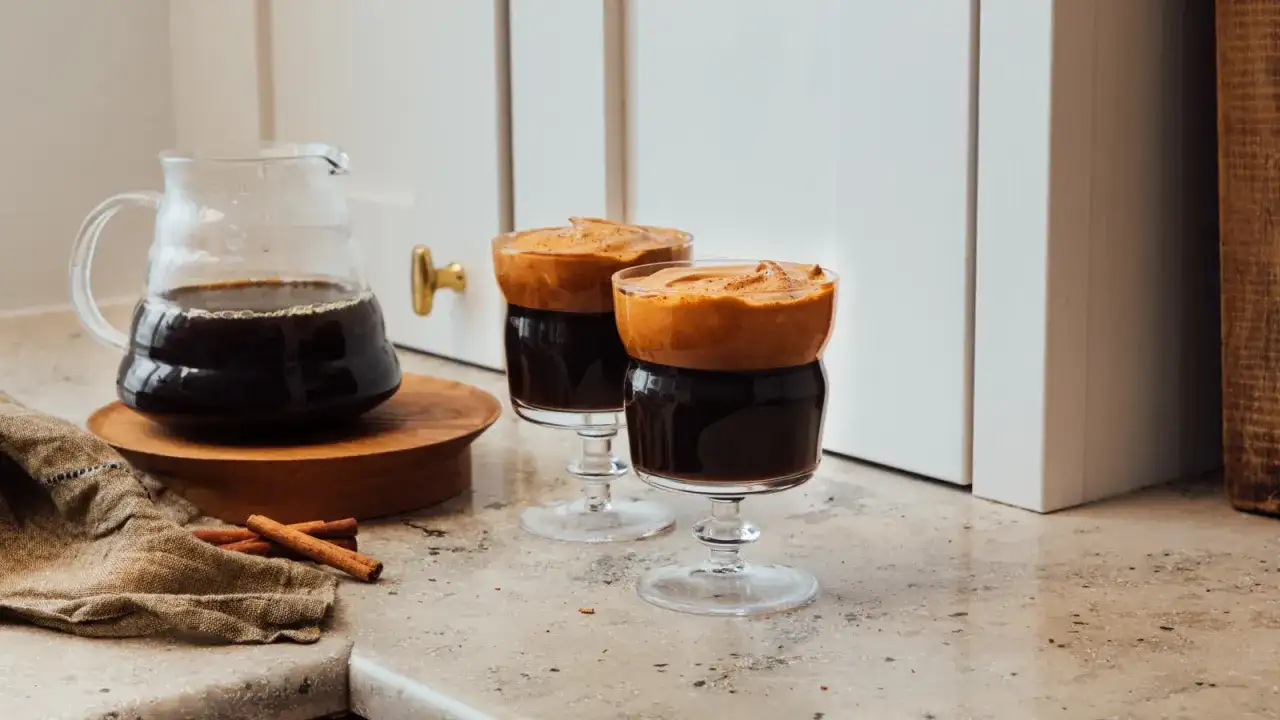 Två glas med mörkt kaffe toppat med fluffigt skum, en härlig dalgona kaffe-upplevelse. Bredvid står en karaff med kaffe och kanelstänger.