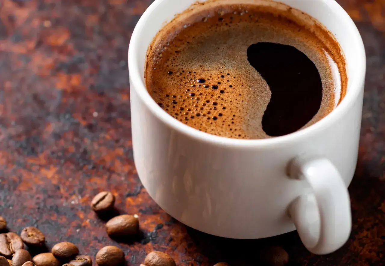 En kopp varmt kaffe med skummig yta, omgiven av kaffebönor. Perfekt för att känna att blir man pigg av kaffe.