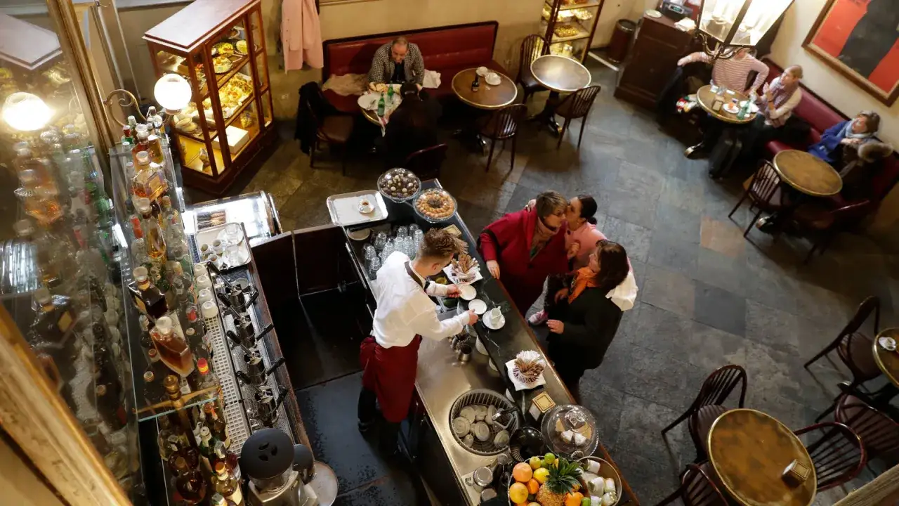 En livlig scen p&aring; ett kaf&eacute; i Italien. En barista f&ouml;rbereder kaffe bakom disken medan g&auml;ster sitter vid borden och njuter av sina drycker och bakverk.