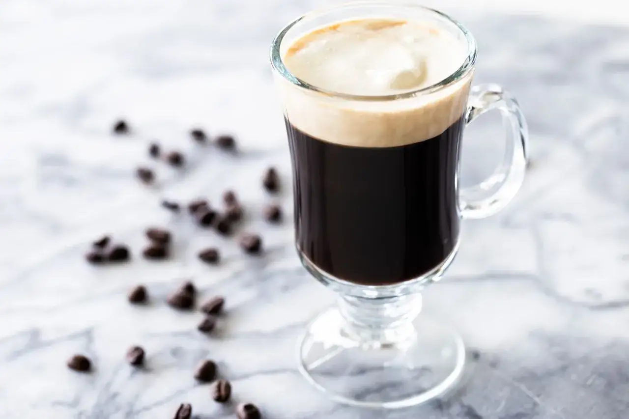 En Irish coffee med kr&auml;migt skum, en hyllning till dess historia. Kaffeb&ouml;nor sprider sig p&aring; marmorbordet.