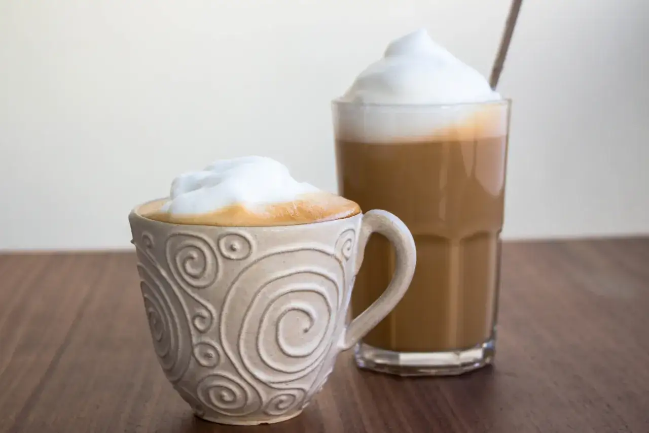 Tv&aring; kaffedrycker, en cappuccino i en m&ouml;nstrad kopp och en latte i ett glas, st&aring;r p&aring; ett tr&auml;bord.