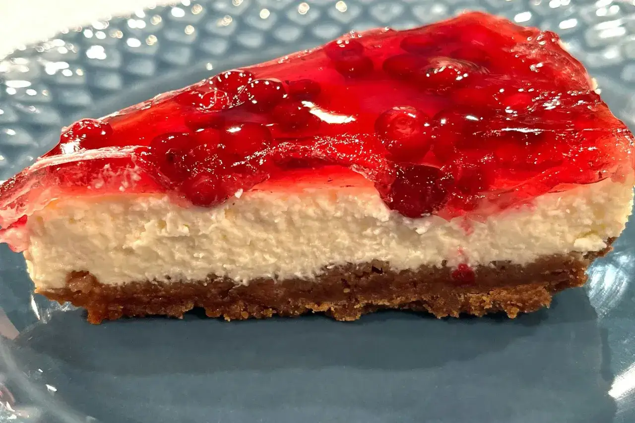 En bit kr&auml;mig saffranscheesecake glutenfri med r&ouml;tt gel&eacute;lager och b&auml;r, serverad p&aring; en bl&aring; tallrik.