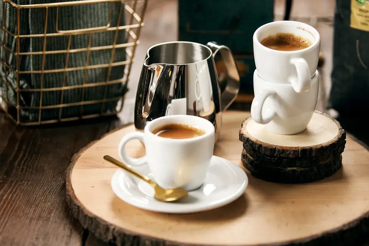Två koppar espresso ristretto, en med en guldig sked, står på ett träfat. En mjölkkanna och en korg med en filt i bakgrunden.