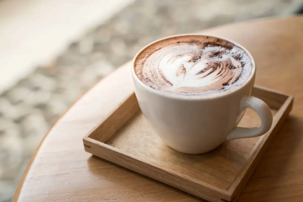 En vit kopp med ett hj&auml;rtm&ouml;nster i skummet, serverad p&aring; en tr&auml;bricka. En perfekt start p&aring; dagen med ett spanskt kaffe.