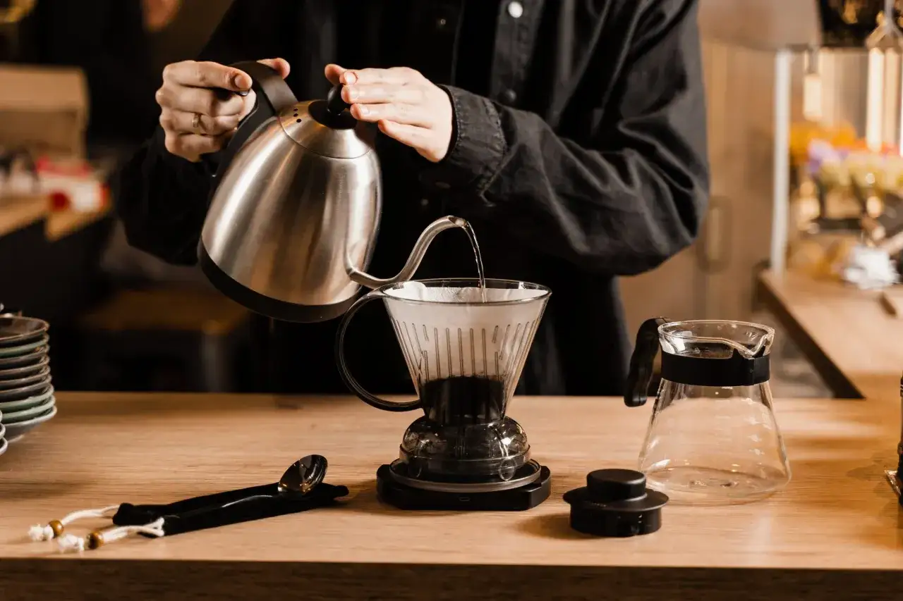 Person h&auml;ller vatten fr&aring;n en silverf&auml;rgad vattenkokare &ouml;ver kaffepulver i en glasfilterh&aring;llare. En karaff st&aring;r bredvid. Perfekt f&ouml;r att m&auml;ta hur mycket kaffe per liter vatten.
