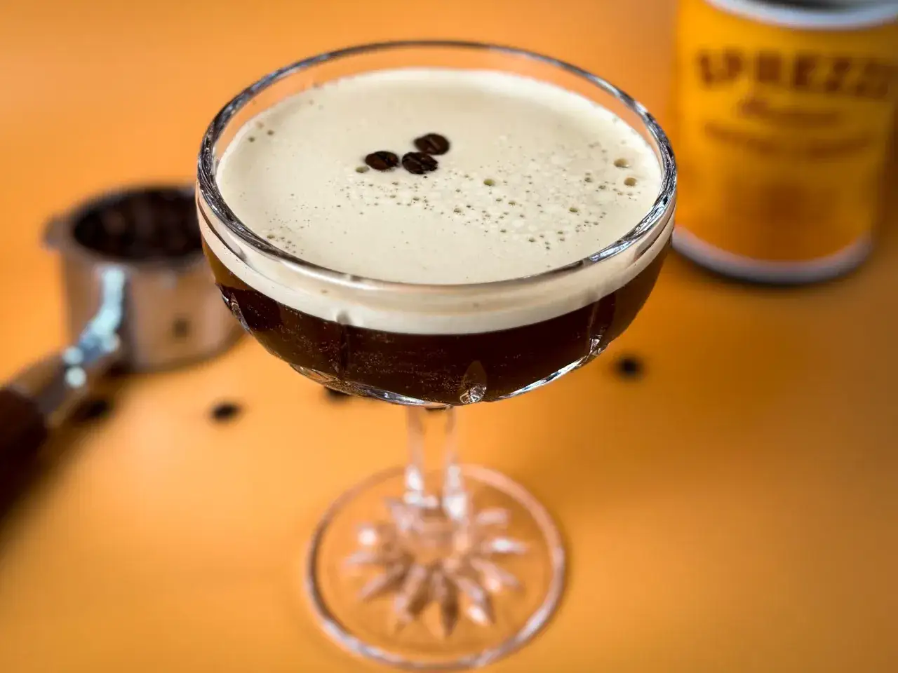 En lyxig espresso martini med kr&auml;mig skumtopp, dekorerad med kaffeb&ouml;nor. Bredvid syns en portafilter med bryggkaffe.