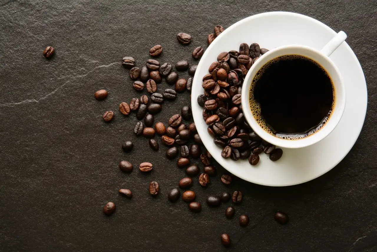 En kopp med mörkt java kaffe står på en vit tefat, omgiven av rostade kaffebönor på en mörk yta.