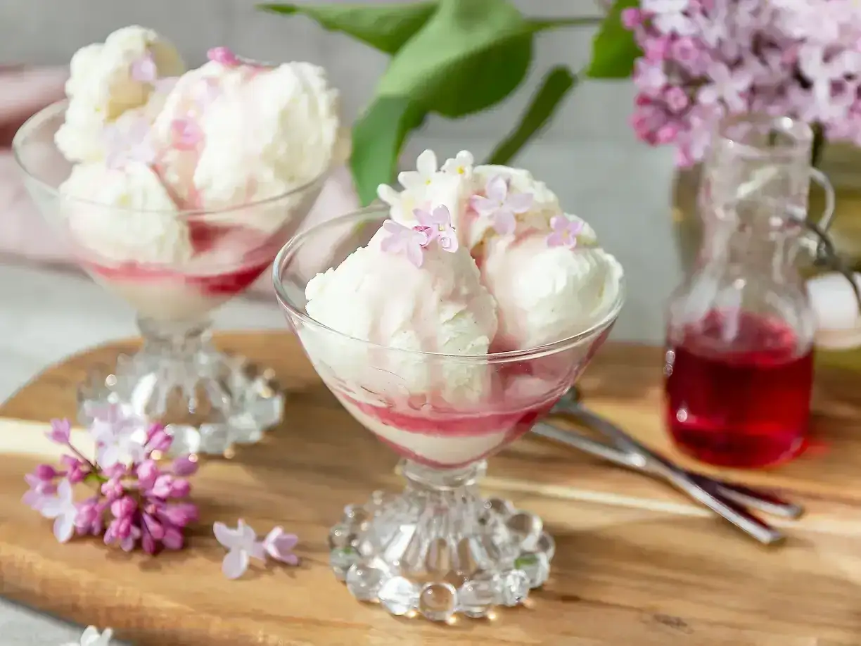 Glassbägare med vaniljglass, toppad med syrenblommor och röd sirap. En flaska med sirap och syrenkvistar i bakgrunden. Ett härligt syrensirap recept!