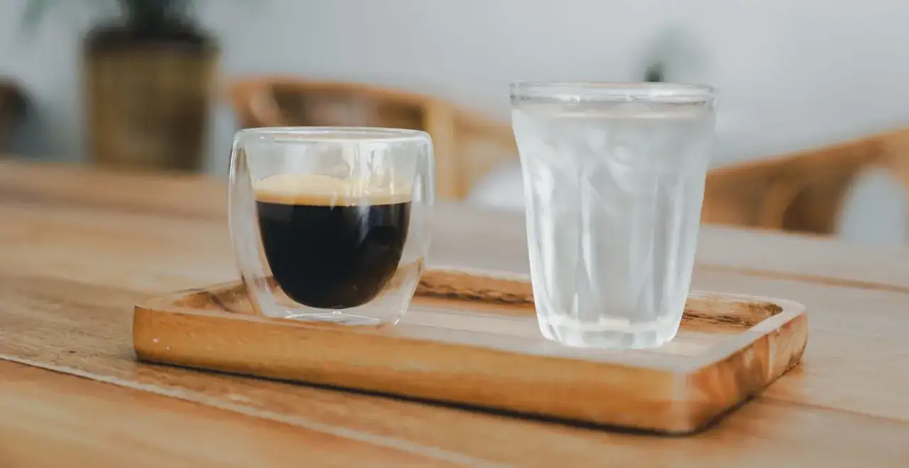 En dubbelv&auml;ggig glasbeh&aring;llare med americano kaffe och ett glas vatten p&aring; en tr&auml;bricka.