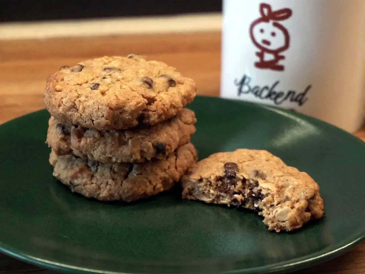 En hög med gammaldags havrekakor med chokladbitar på en grön tallrik. En kaka är uppäten. En mugg med en kaninfigur och texten