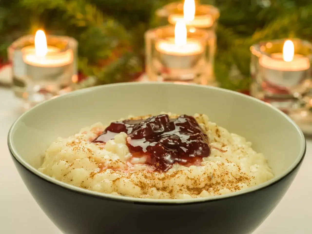 En skål med krämig risgrynsgröt, toppad med sylt och kanel. Tända ljus och granris skapar en mysig julstämning, perfekt för ett risgrynspudding recept.