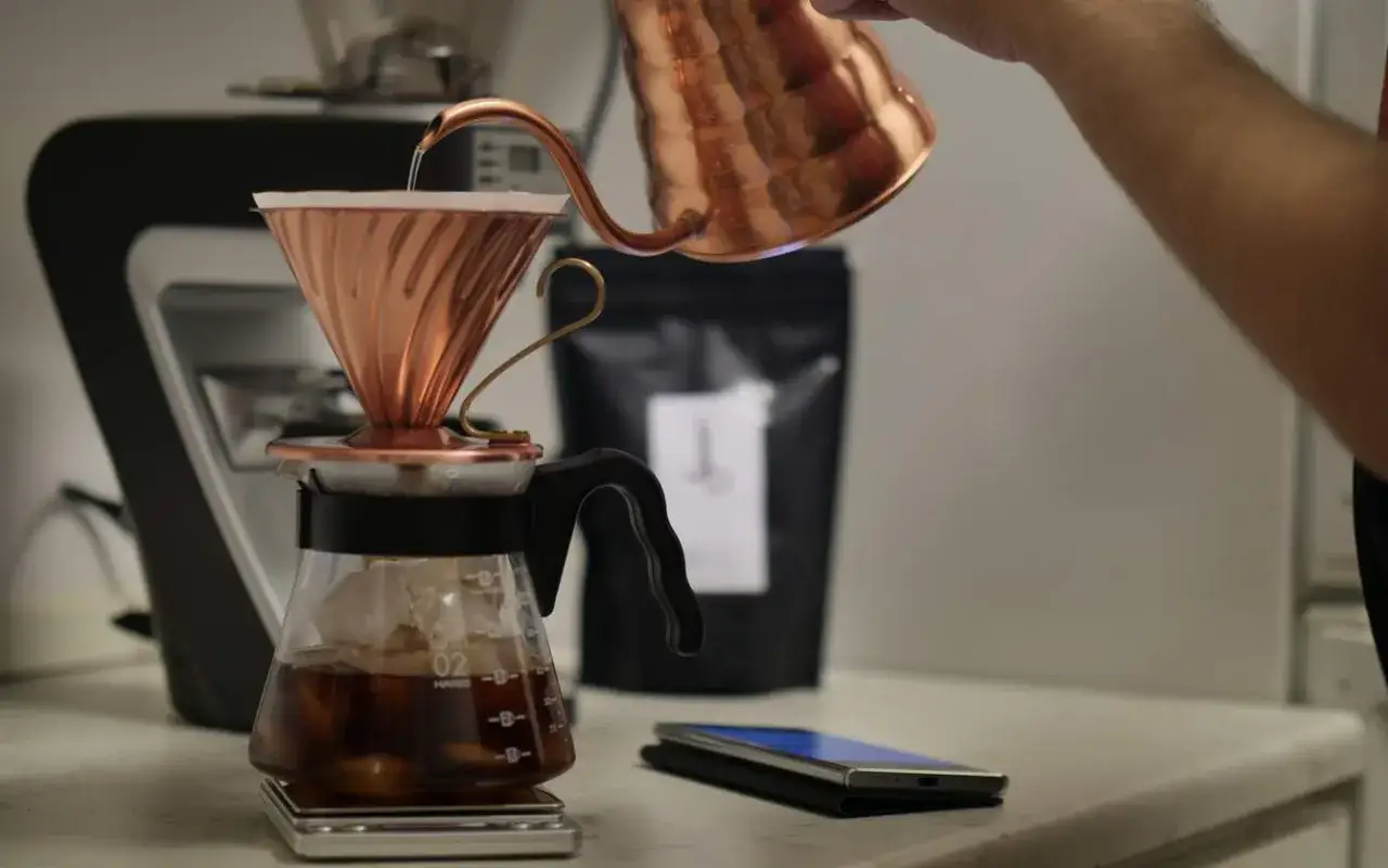 En hand h&auml;ller varmt vatten &ouml;ver kaffefilter i en kopparkopp, vilket skapar en uts&ouml;kt espresso i kaffebryggaren nedanf&ouml;r.