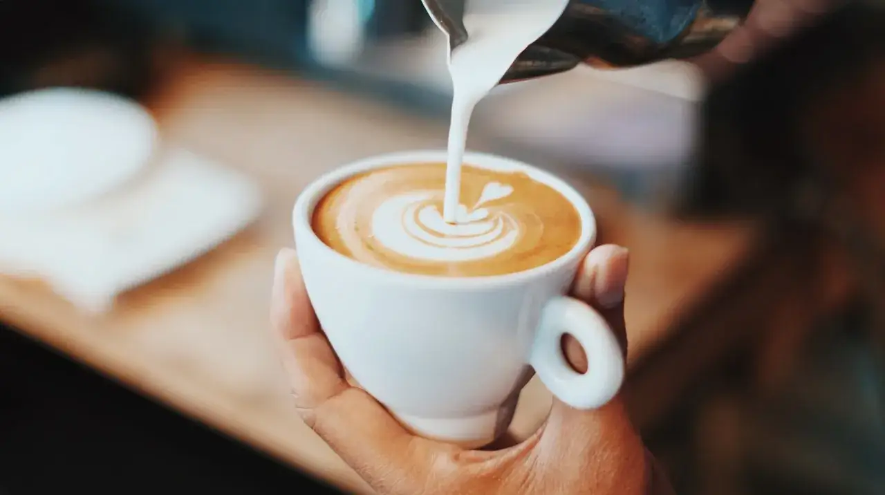 En hand h&aring;ller en vit kopp med en vacker kaffe latte, d&auml;r mj&ouml;lken skapar ett hj&auml;rta i skummet.