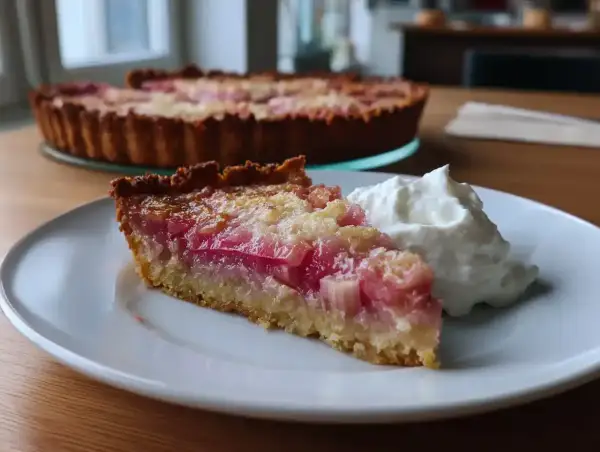 En saftig bit glutenfri rabarberpaj med en fluffig klick grädde. Hela pajen syns i bakgrunden.