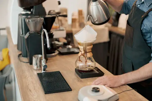 Barista häller vatten över kaffefilter i en Chemex. En kaffekvarn och en kran syns i bakgrunden. Intressant kaffe fakta!