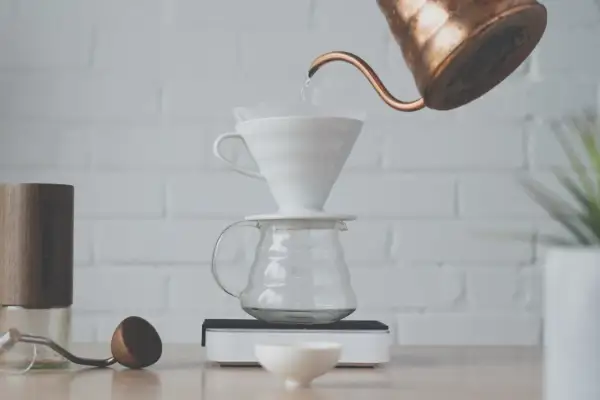 Vatten hälls från en kopparkanna över ett filter i en vit keramiktratt, som står på en glaskaraff. En kaffekvarn och en träsked ligger bredvid. Det är dags att brygg.