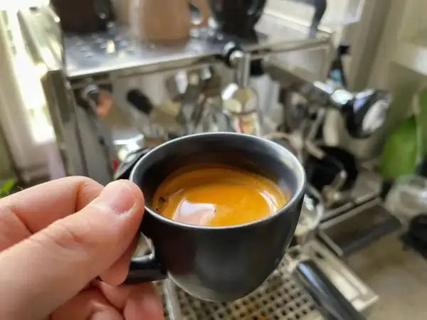 En hand håller en kopp med gyllene spanskt kaffe framför en espressomaskin.