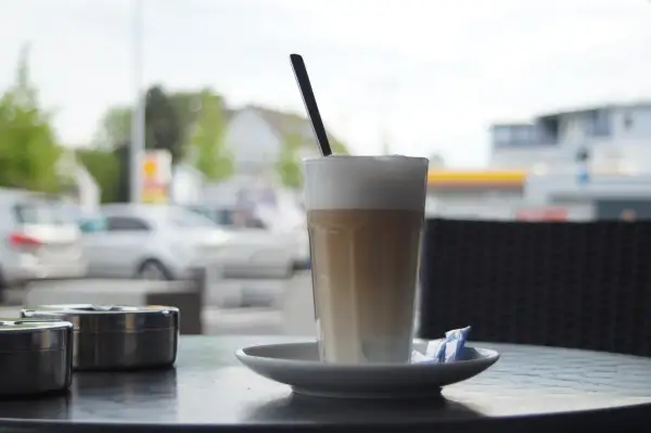 En hög latte macchiato med skummjölk och en sked på ett fat, bredvid två askkoppar.