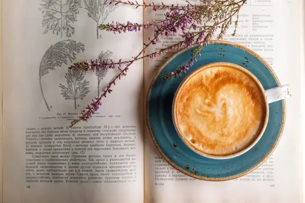 En kopp kaffe med lattekonst vilar på en öppen bok med botaniska illustrationer. En lila blomsterkvist ligger över boken och koppen, som om den vore en del av en studie för att inte frysa kaffe.
